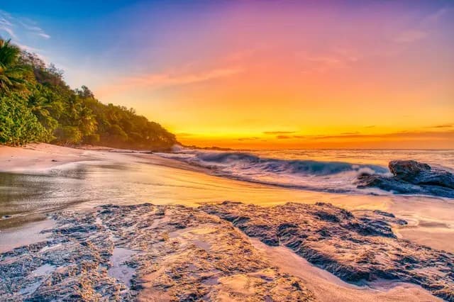 A vibrant sunset casts colorful reflections on a sandy beach with gentle waves and rocky formations.