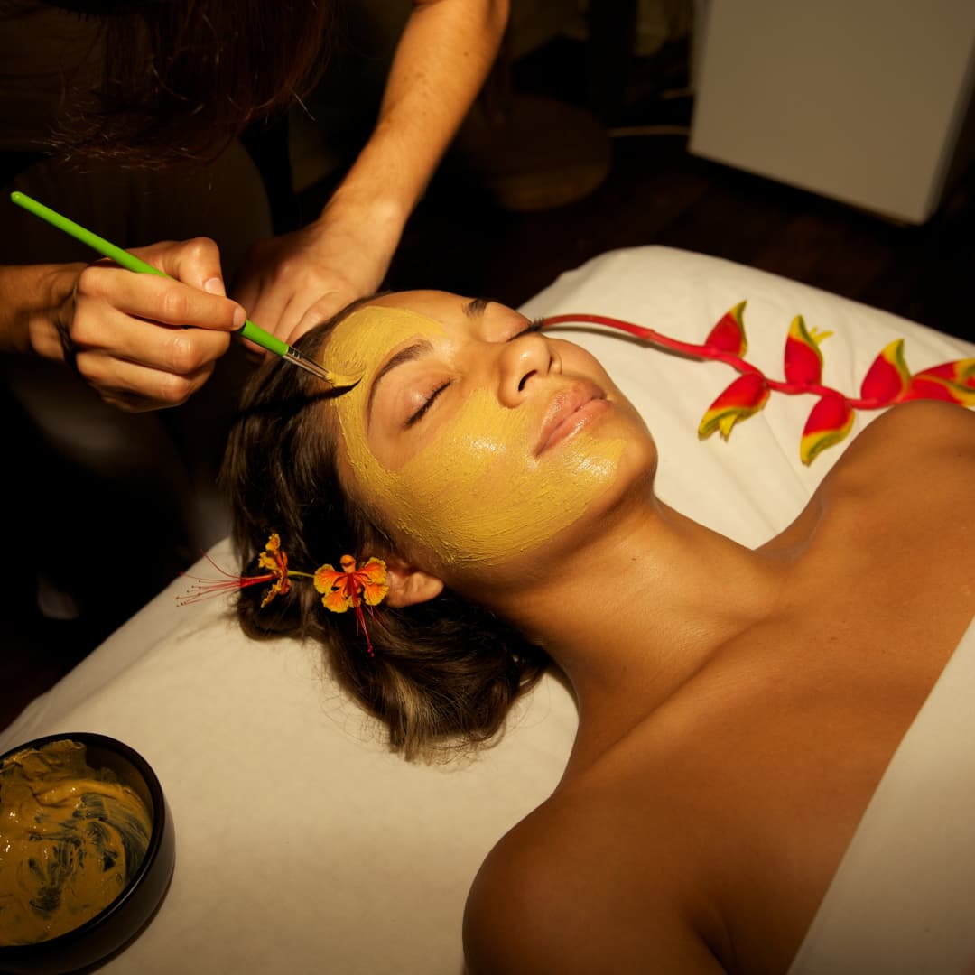 A woman draped in a sheet lying face up looking blissful with her eyes closed while her face is being brushed with a creamy facial treatment