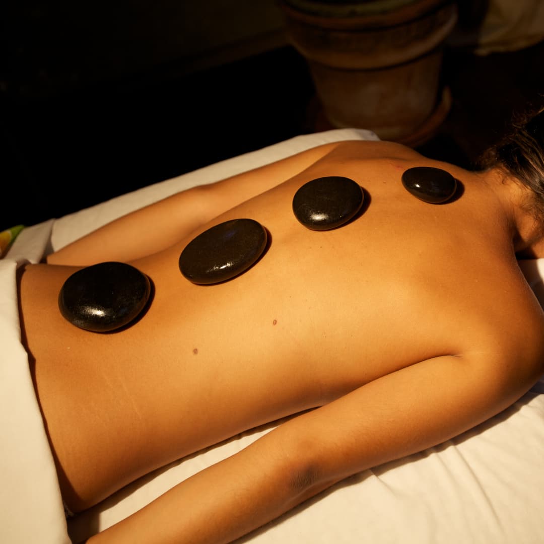 A woman lying face down on a massage table draped in a sheet around her waist with shiny, smooth round stones placed down her spine