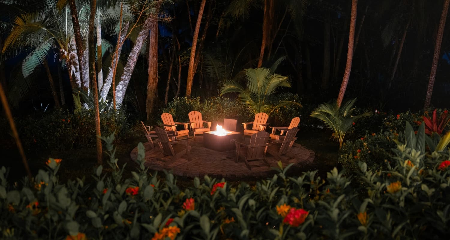 A cozy outdoor seating area with wooden chairs surrounding a fire pit, illuminated at night amidst tropical foliage.