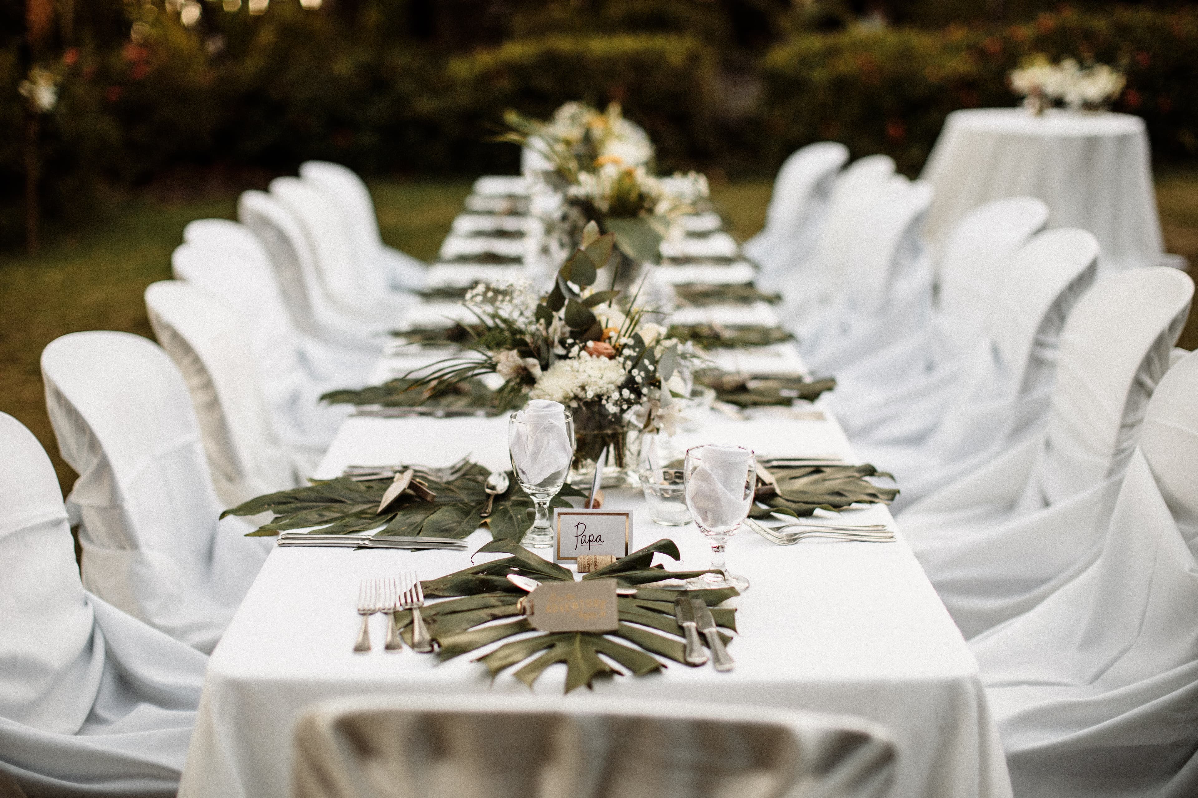 A long dining table set up for an elegant, outdoor reception with white tablecloth, white draped chairs, tropical floral centerpieces, and placecards