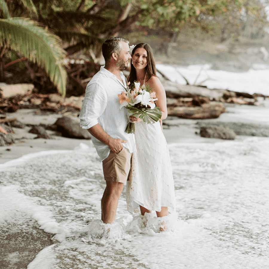 A couple stands in shallow water on a beach, smiling and holding a bouquet of flowers.
