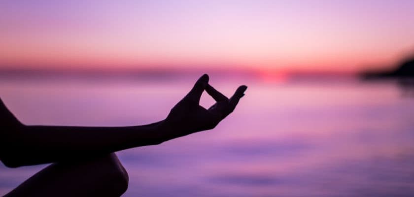 A silhouetted hand forms a mudra against a serene sunset over water.