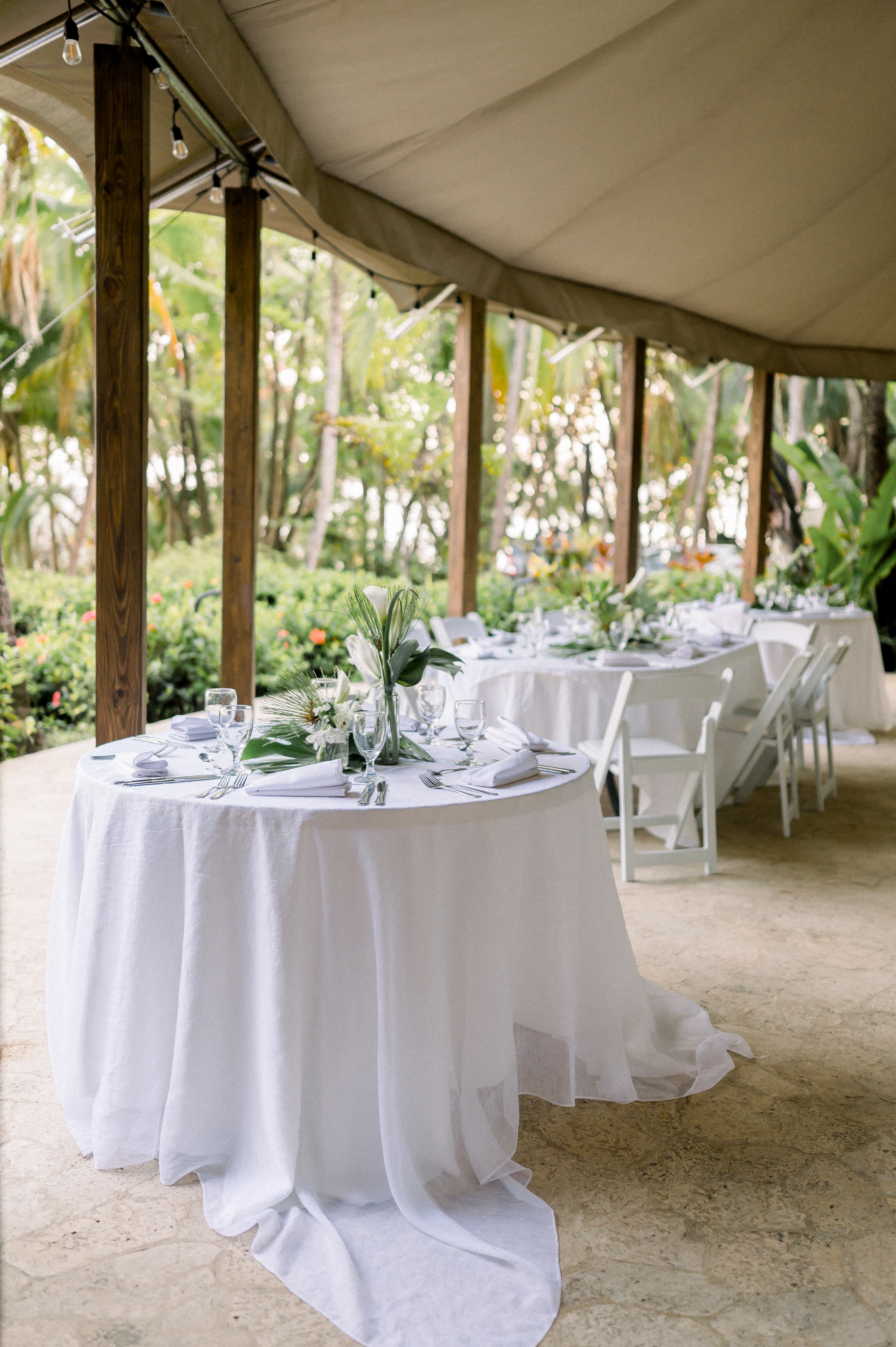 A large round wooden gazebo set up for a wedding reception with round tables set with long, flowing white tablecloths, white chairs and tropical floral centerpieces