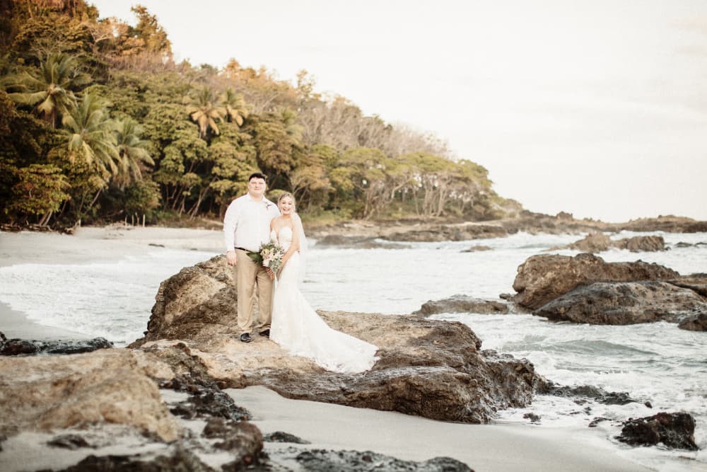 A smiling wedding couple standing on large rocks with ocean waves and palm trees and other trees around them.