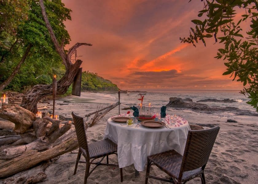 A romantic dinner setup on a beach at sunset, featuring a decorated table with chairs and surrounded by tropical scenery. A romantic dinner setup on a beach at sunset, featuring a decorated table with chairs and surrounded by tropical scenery.