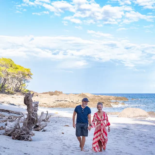 A couple walks hand in hand along a sandy beach under a blue sky.