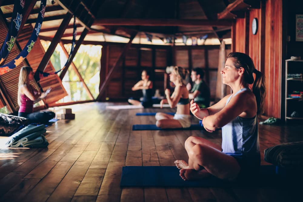 Several people sitting cross-legged on yoga mats with their arms bent in front of them and their hands crossed