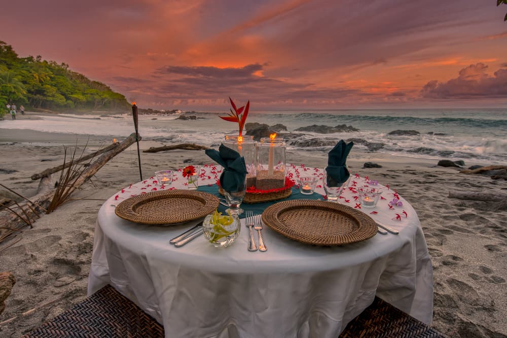 Dining table on beach with table cloth, driftwood and ocean in the background with the sun setting and the jungle off shore