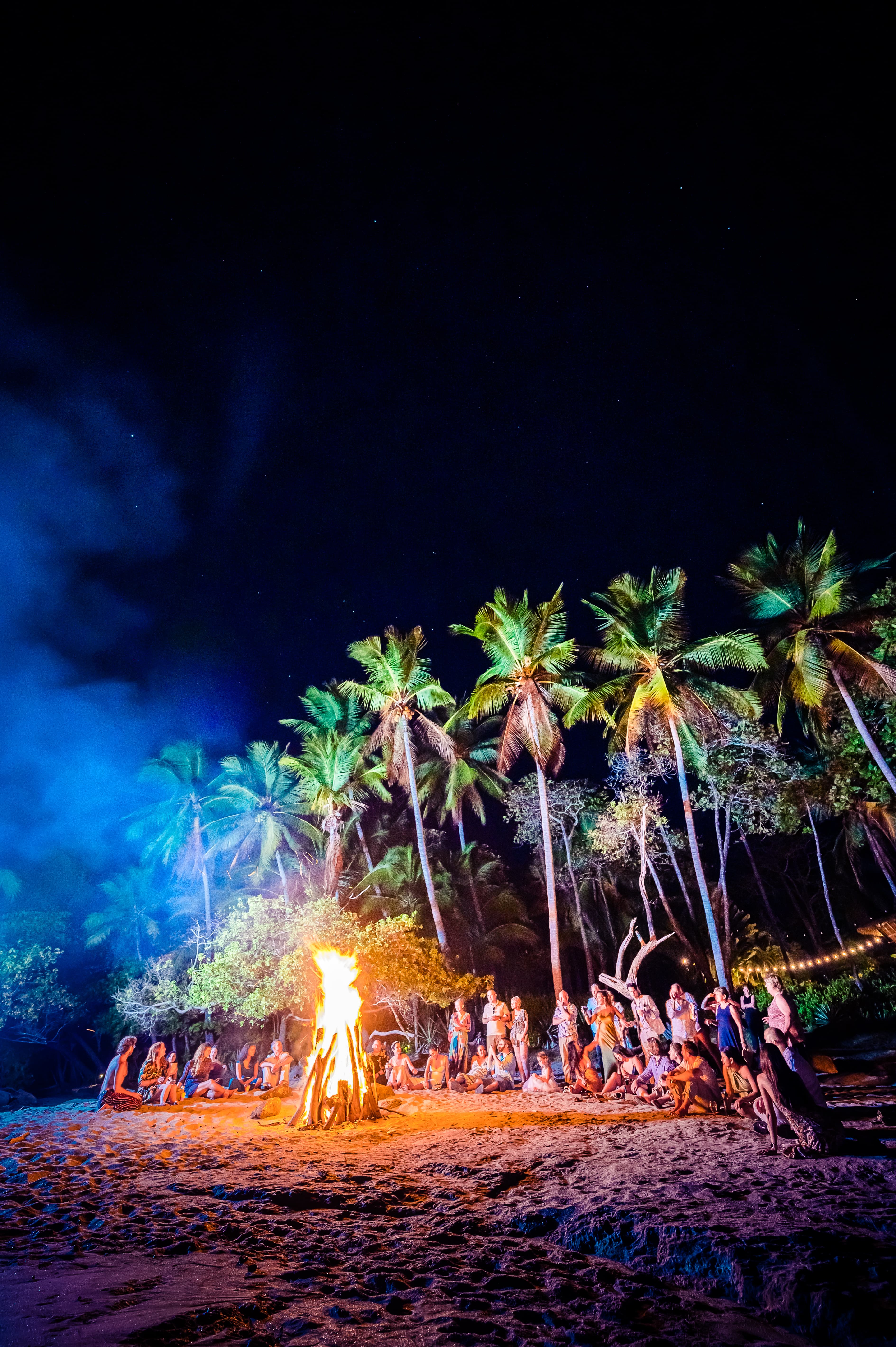 Photo of people all standing and sitting on the beach with a large bonfire in the foreground and the jungle in the background.