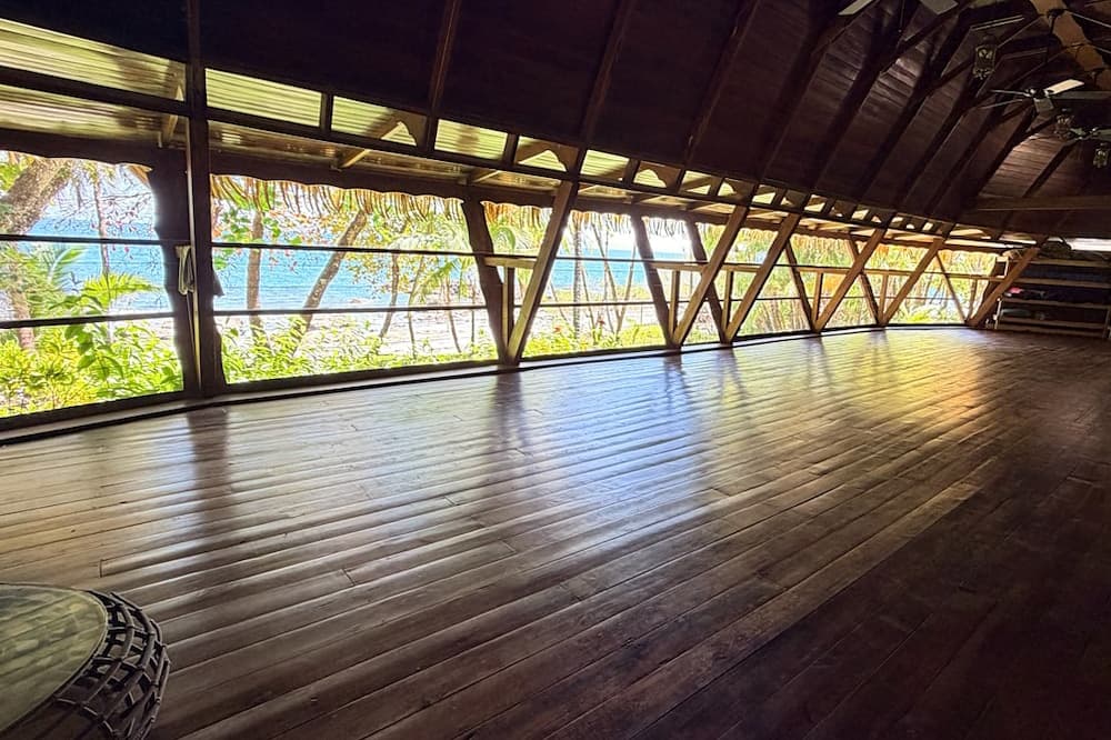 Interior of a wooden room with large windows overlooking a tropical landscape and ocean.
