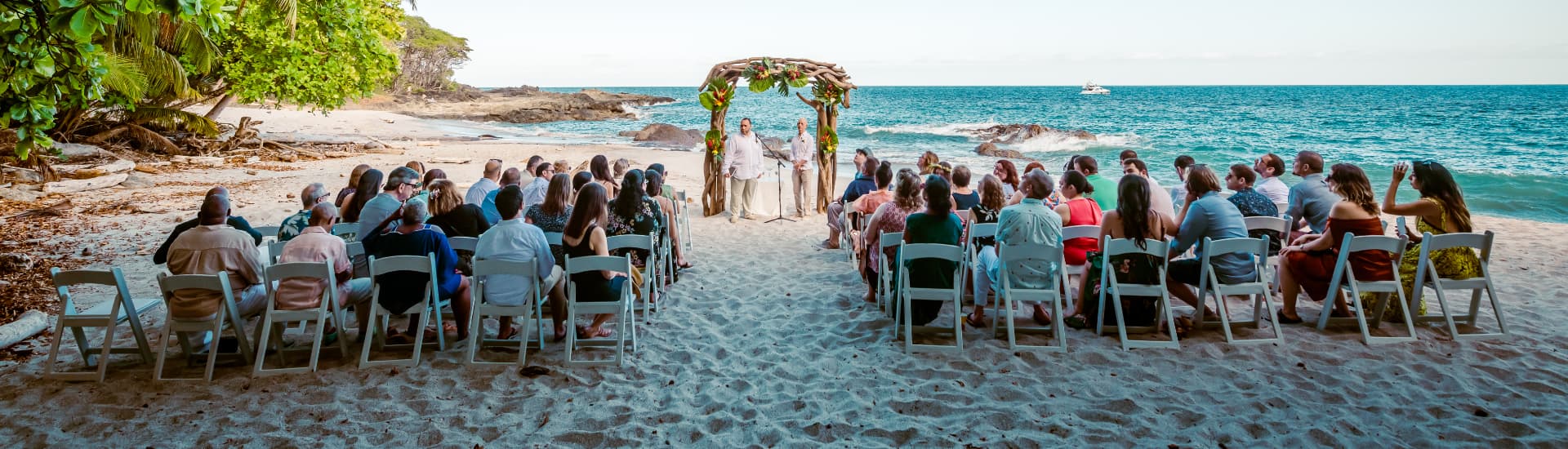 A wedding on a sandy beach with well wishers on each side of a native arch with the groom and an officiant below the arch, and a blue ocean behind them, with a jungle on the other side of the beach.