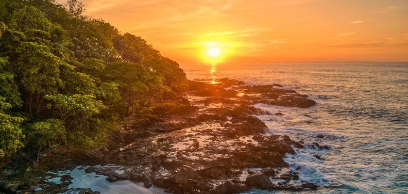 rocky shoreline with waves crashing and trees on the left, under a vibrant sunset over the ocean