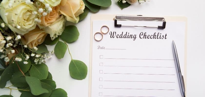 wedding checklist on a clipboard with two rings flowers and greenery on a white table