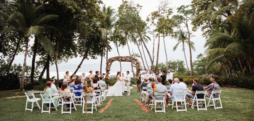 a couple exchanges vows under a wooden arch surrounded by guests on a grassy tropical outdoor setting at ylang ylang beach resort
