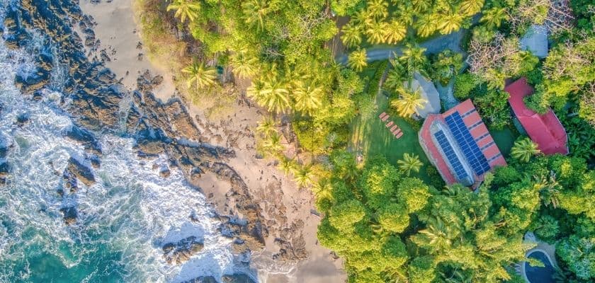 aerial view of a tropical coastline with rocky shores lush green palm trees and a red roofed resort building nestled in the jungle beside the ocean